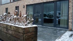 A wooden planter box with snow-covered plants in front of a brick building with large glass doors.