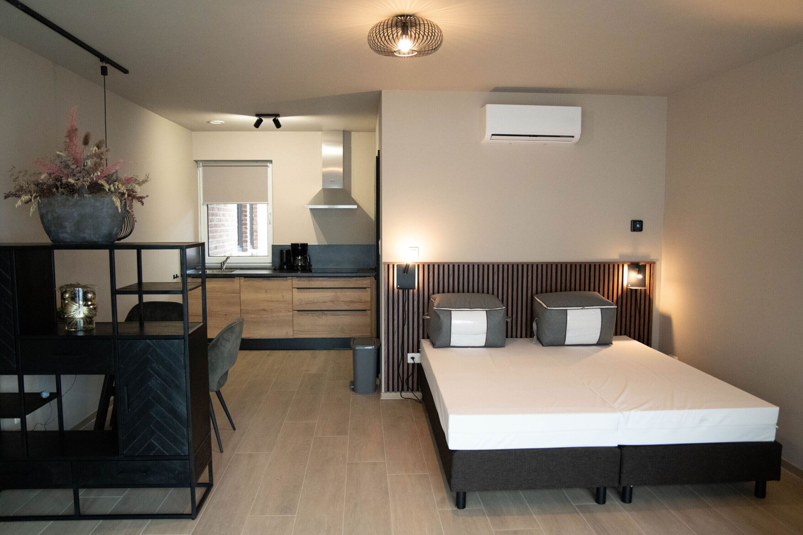 Ein modernes Schlafzimmer mit Bett, Nachttischen und einer kleinen Nachttischkonsole. Es gibt eine Küchenzeile mit Fenster und einer Pendelleuchte.