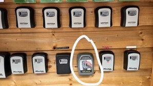 A display of various locks and keys, including brands like Grifema and Master Lock, arranged on a wooden panel with labels indicating different apartment names. - Charming & cosy T2, close to Casino Park and Spa (Argelès-Gazost)