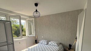 A bedroom with a checkered bedspread, a radiator, a window with a view of greenery, and a hanging light fixture. - Charming & cosy T2, close to Casino Park and Spa (Argelès-Gazost)