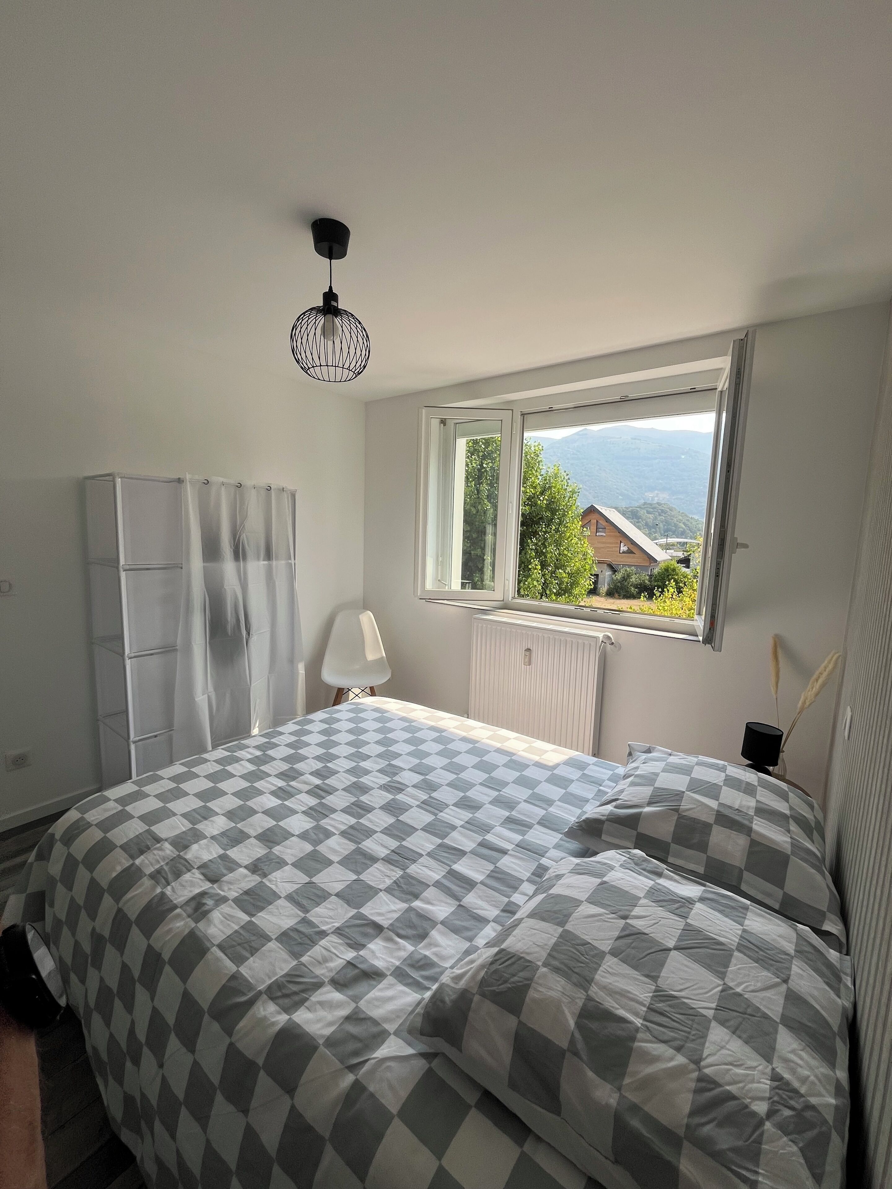 Un dormitorio con colcha a cuadros, un radiador, una ventana que ofrece vistas a la vegetación y montañas, una silla blanca y una lámpara colgante.