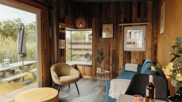 A cozy cabin interior with wooden walls, a dining area with a table and chairs, a sofa, and a hanging light fixture. - Solglimt 2 pers holiday home with stunning views (Callantsoog)