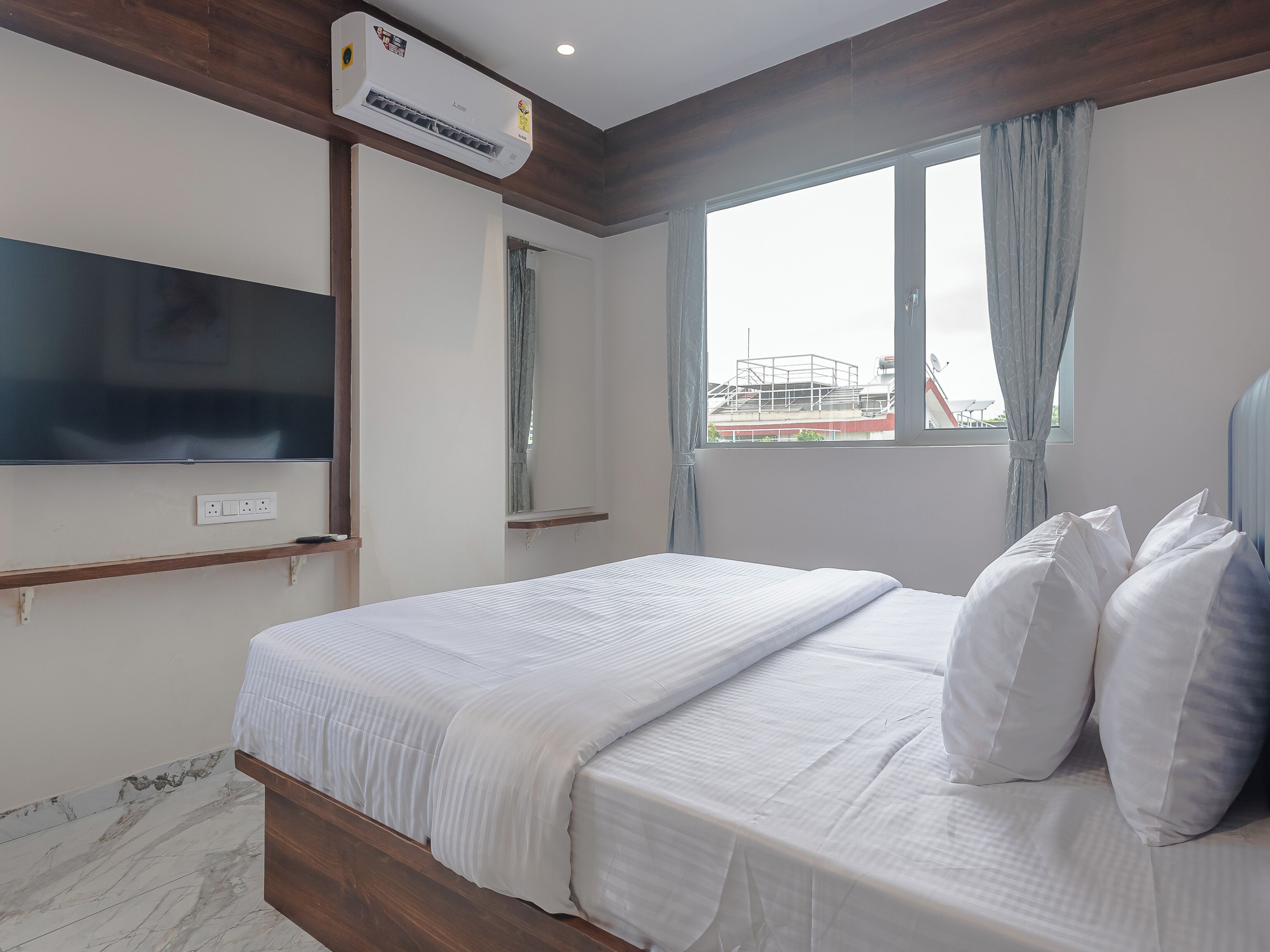 Een hotelkamer met een bed, een wandtelevisie, een airconditioning en een raam met gordijnen, met uitzicht op een buitenbouw.