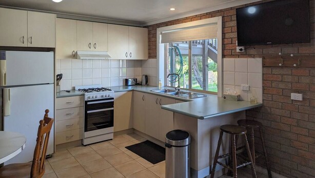 Cozinha moderna com armários brancos, fogão a gás, forno embutido, janela com vista e TV fixada na parede de tijolos.