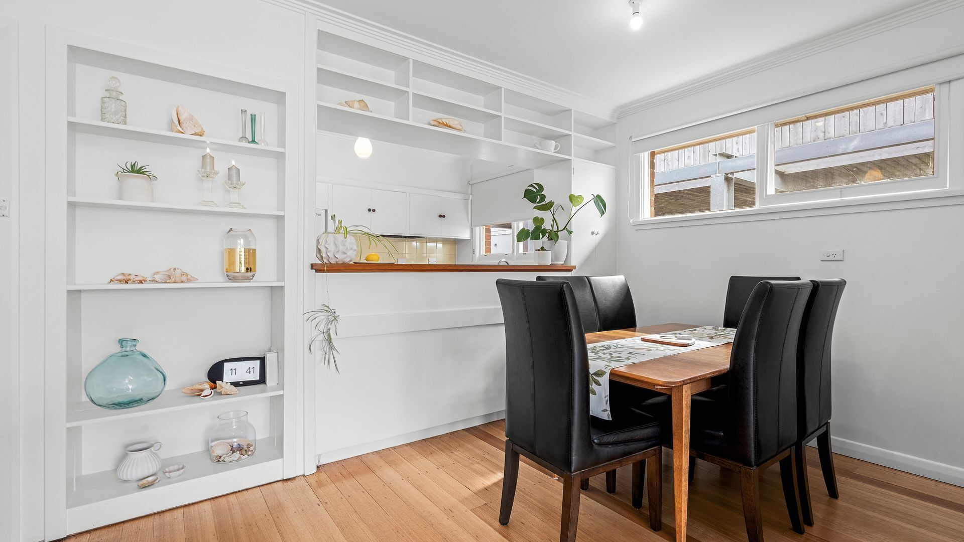 A dining area with a wooden table and black chairs, white shelving with decorative items, and a kitchen area with a window.