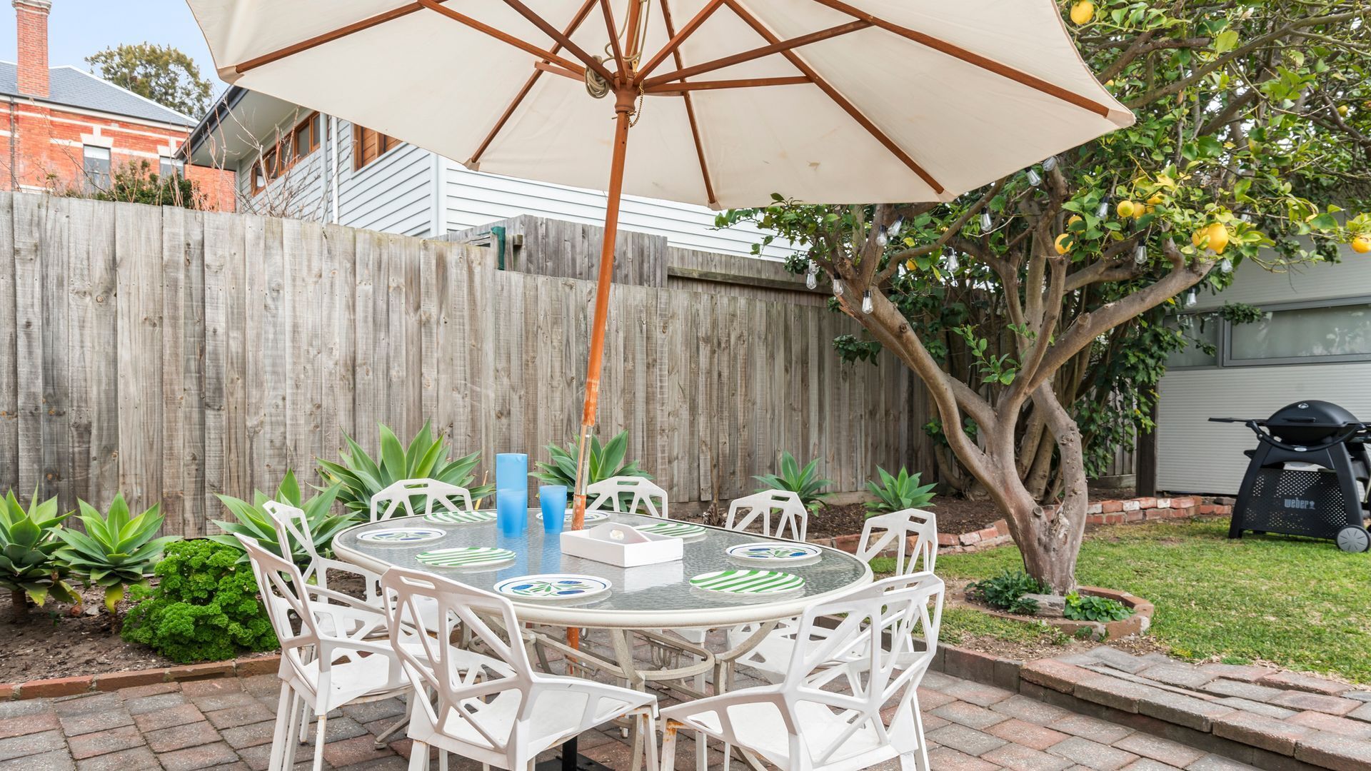 A patio with a round table, white chairs, a large umbrella, and a wooden fence in the background.