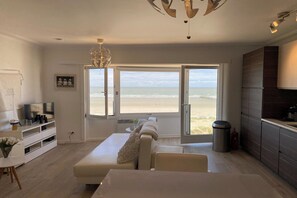 A modern living room with a large window overlooking a beach, a white sofa, a TV, and a minimalist kitchen. - Sea-view apartment in Nieuwpoort (NIEUWPOORT)