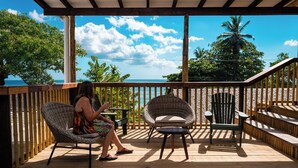 A wooden deck with a woman sitting on a wicker chair, another chair, and a small table. - Beach House Beau Vallon (Mahe Island)