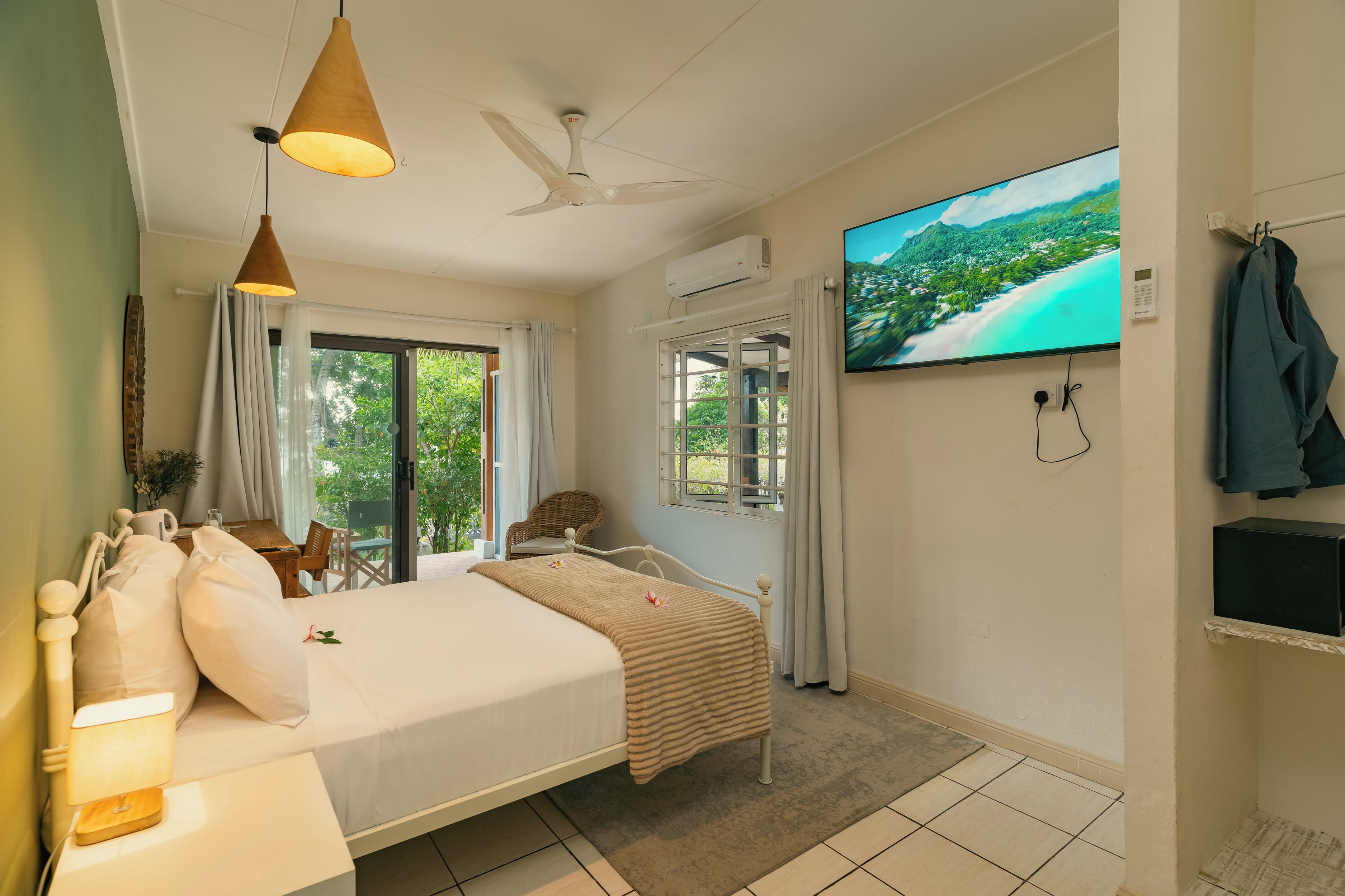Ein Hotelzimmer mit Bett, Fernseher, Deckenventilator und Blick auf eine tropische Landschaft.
