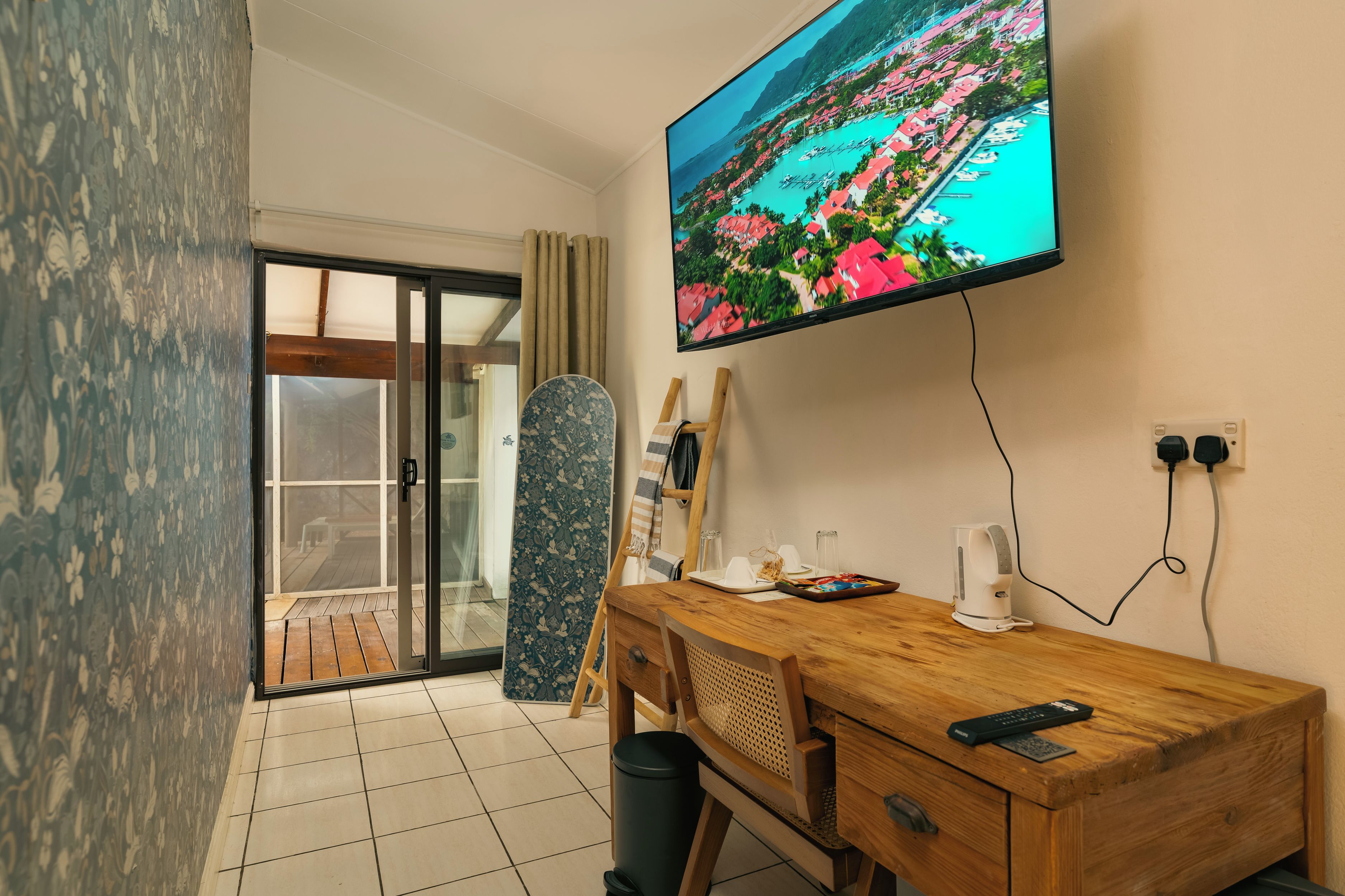 A hotel room with a TV displaying a scenic view, a wooden desk with a remote control, a patterned curtain, and a sliding glass door leading to a balcony.