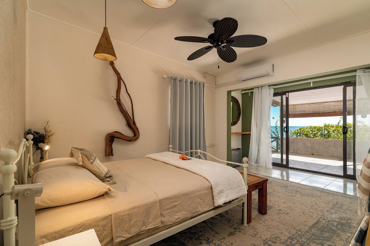 Een slaapkamer met een bed, een plafondventilator, een hanglamp, uitzicht op het omliggende landschap en een schuifdeur.'