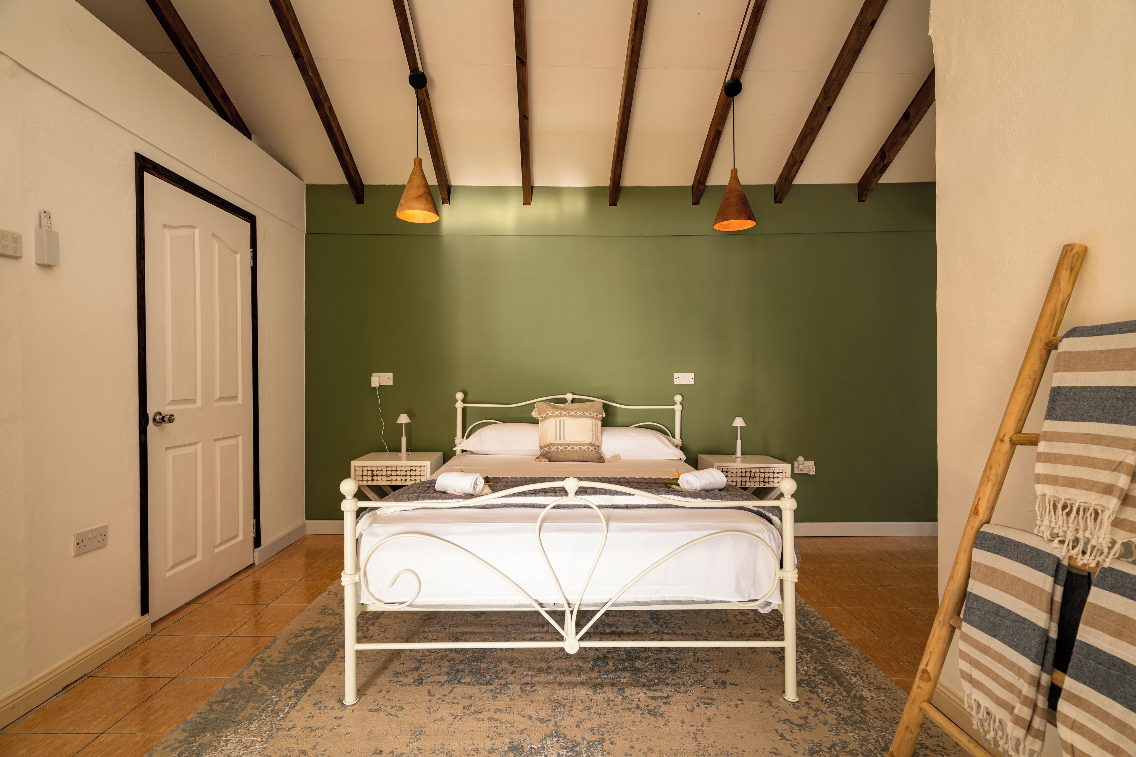 Un dormitorio con estructura de cama metálica blanca, paredes verdes y techo de madera. Hay dos mesitas de noche con lámparas, una puerta y una manta a rayas sobre un atril de madera.