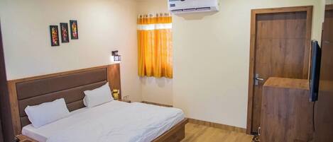 A hotel room with a bed, a ceiling fan, an air conditioning unit, a wooden wardrobe, and a window with curtains.