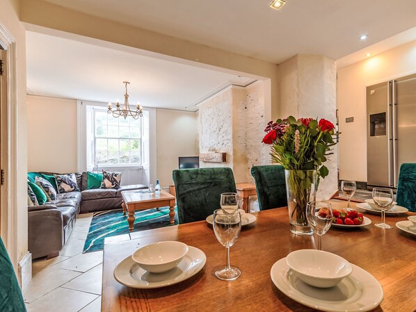 Comedor con una mesa de madera dispuesta para cuatro personas, rodeada de sillas tapizadas en verde. Un jarrón con flores rojas está sobre la mesa, y hay platos, vasos y un bol.