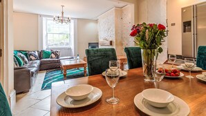 Comedor con una mesa de madera dispuesta para cuatro personas, rodeada de sillas tapizadas en verde. Un jarrón con flores rojas está sobre la mesa, y hay platos, vasos y un bol.