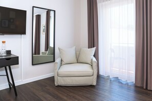 Habitación de hotel con sofá, escritorio, botella, vasos, espejo y ventana con cortinas.
