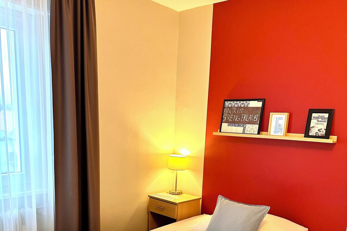 A hotel room with a bed, a nightstand with a lamp, and a red accent wall with framed pictures.
