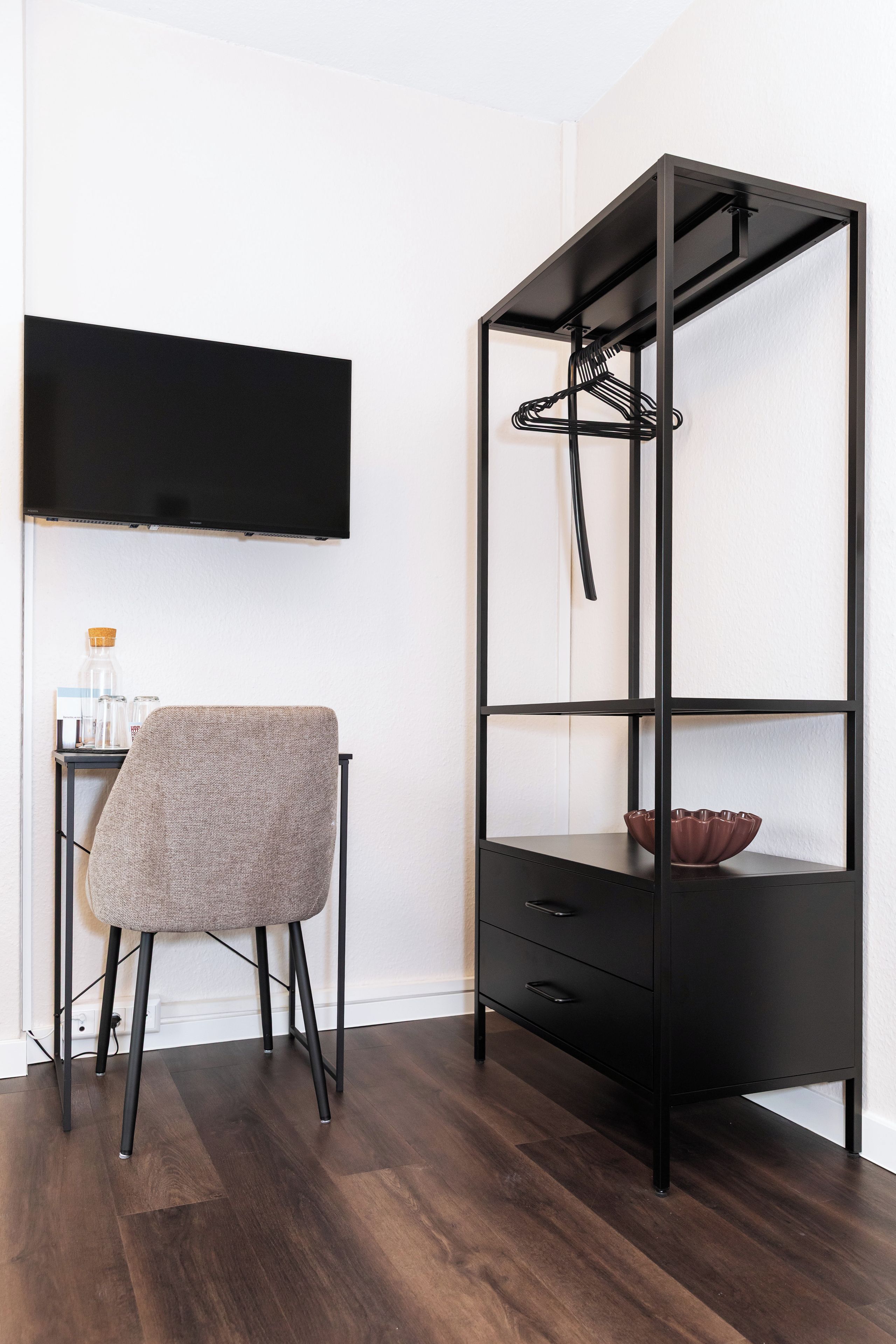 Een moderne kamer met een zwarte metalen schap, een grijs gestoffeerde stoel en een flatscreen televisie aan de muur.