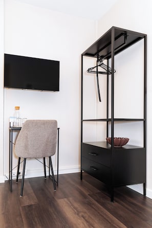A modern room with a black metal shelving unit, a grey upholstered chair, and a flat-screen TV mounted on the wall. - Hotel Friedrichstraße  (Lampertheim)