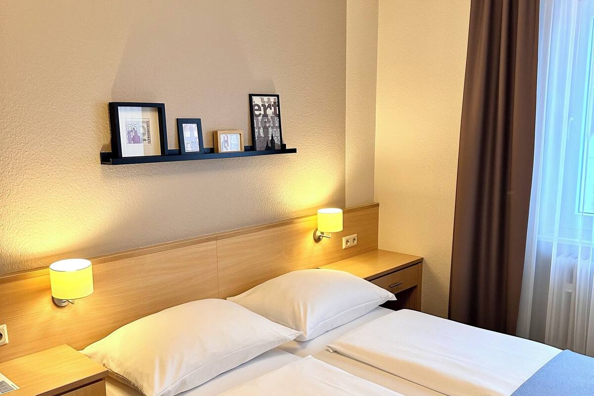 A hotel room with a neatly made bed, two bedside tables with lamps, a shelf with framed photos, and a window with curtains.