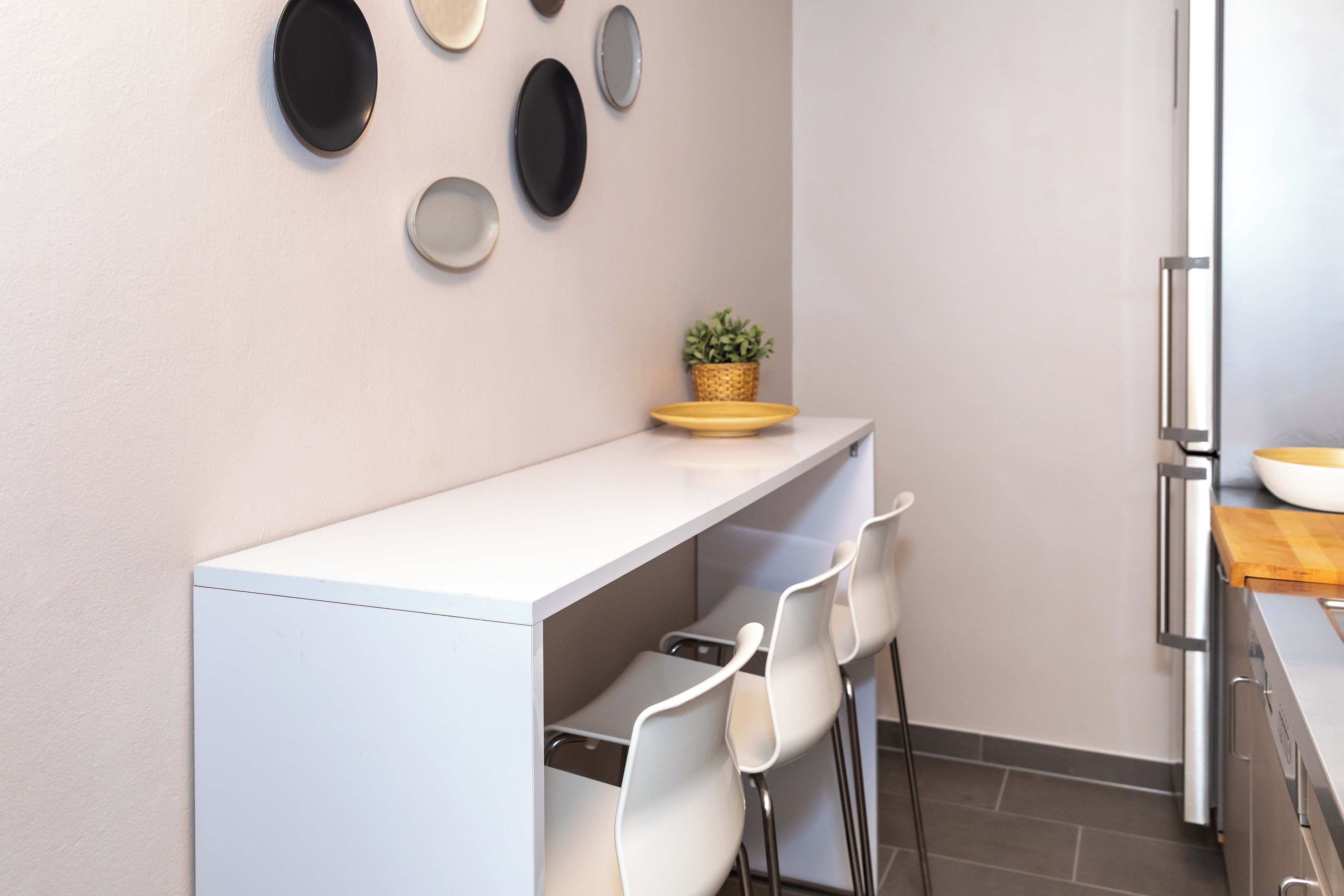 Eine moderne Küche mit einer weißen Arbeitsplatte, zwei weißen Stühlen, einem kleinen Blumentopf und einem Edelstahlkühlschrank.