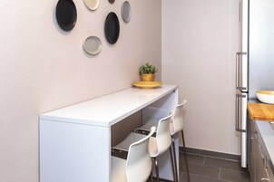 Una cocina moderna con encimera blanca, dos sillas blancas, una pequeña planta en maceta y un refrigerador de acero inoxidable.