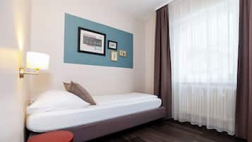 A single bed with a headboard, a bedside table, a wall-mounted lamp, and a window with sheer curtains.