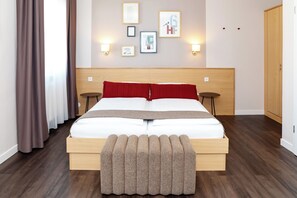 A hotel room with a wooden bed, two bedside tables, a bench at the foot of the bed, and a wall with framed pictures and wall-mounted lights. - Hotel Friedrichstraße  (Lampertheim)