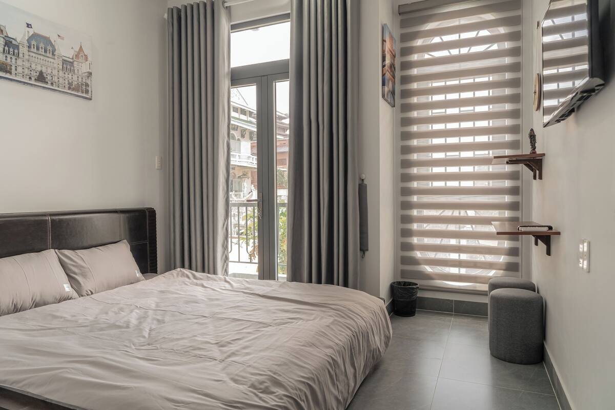 Ein Schlafzimmer mit Bett, Vorhängen, einem Fenster mit Jalousien, einem kleinen Tisch und einem Mülleimer.