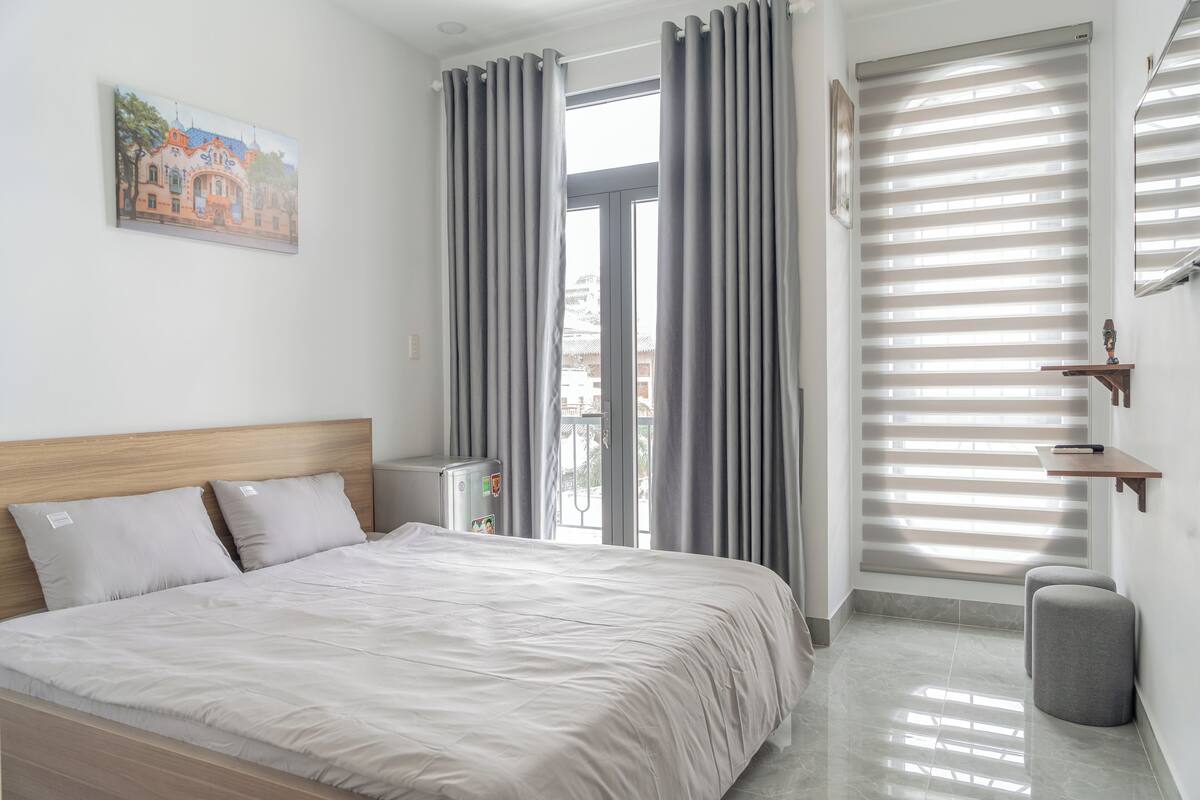 Ein modernes Hotelzimmer mit einem großen Bett, grauen Vorhängen, einem kleinen Tisch und einem Fenster mit Jalousien.