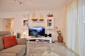 Un salón con una televisión de pantalla plana, un sofá, una mesa de centro y elementos decorativos, incluyendo maquetas de barcos y un jarrón.