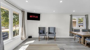 A room with a TV displaying 'NETFLIX,' two chairs, a dining table with chairs, large windows, and a patterned rug. - Spacious home with 6 Beds (Brampton)