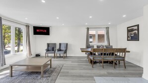 A spacious room with a wooden dining table, two grey armchairs, a TV mounted on the wall, and large windows. - Spacious home with 6 Beds (Brampton)