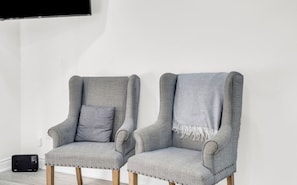 Duas poltronas cinzentas com pés de madeira e um encosto, colocadas em frente a uma parede branca com uma televisão de tela plana fixada acima.