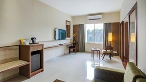 Et hotelværelse med en træsofa, et lille bord, et tv ophængt på væggen, et skrivebord med stol og et vindue med gardiner.