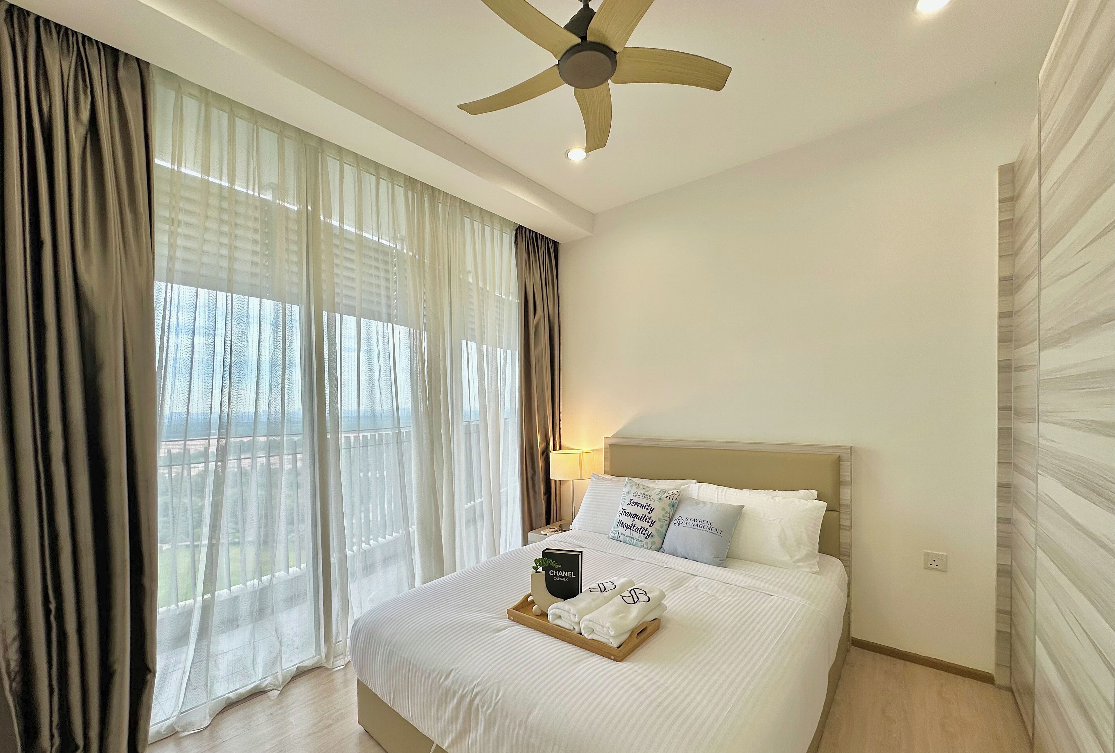 Une chambre d’hôtel avec un lit, un ventilateur de plafond, une fenêtre avec des rideaux et une table de chevet avec une lampe.