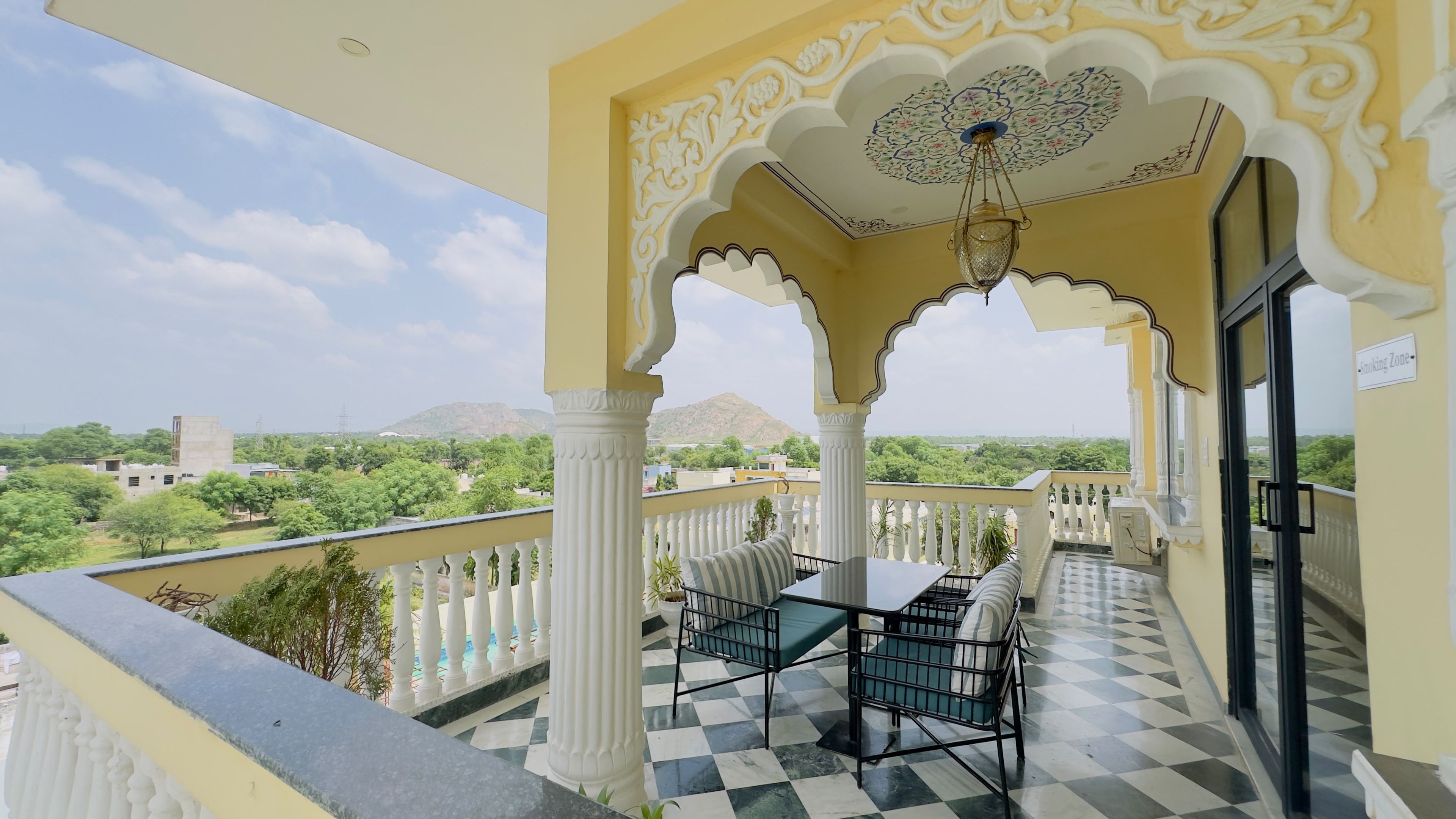 A balcony with a checkered floor, a table with chairs, and a view of greenery and distant hills.
