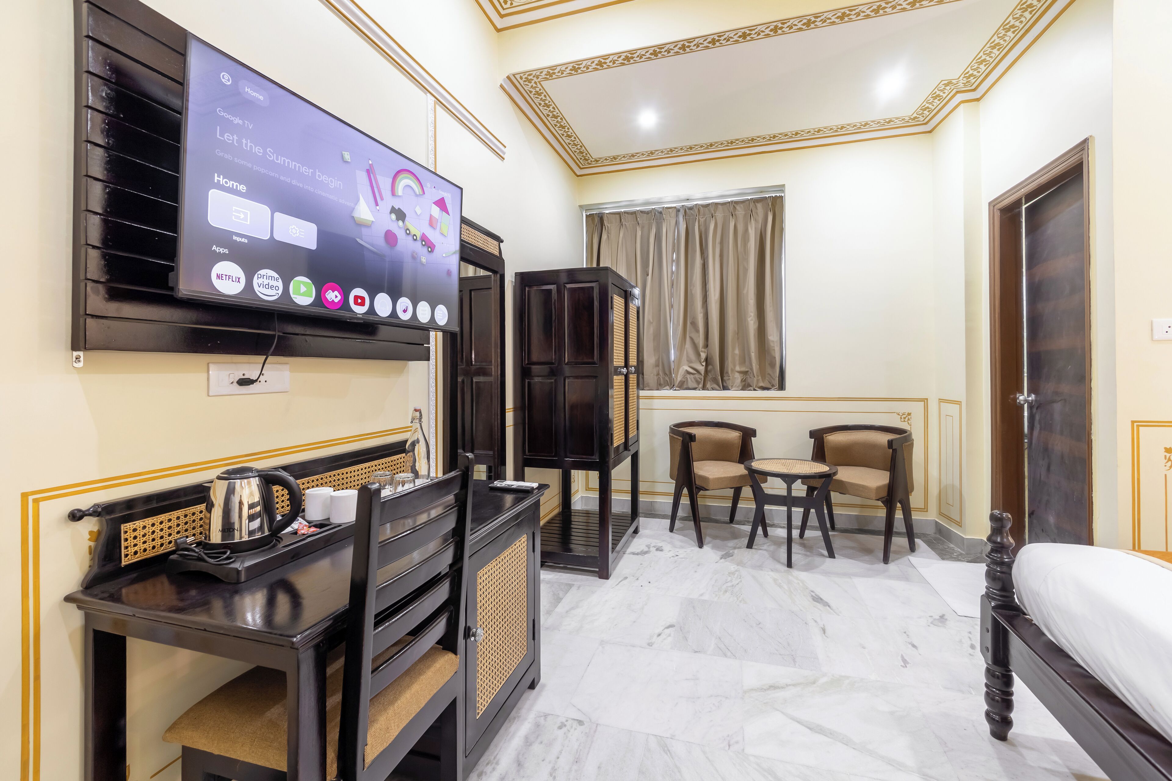 Habitación de hotel con televisor de pantalla plana montado en la pared, una mesita con hervidor y tazas, dos sillas, un mueble con puerta de mimbre y una cama con colchón blanco.
