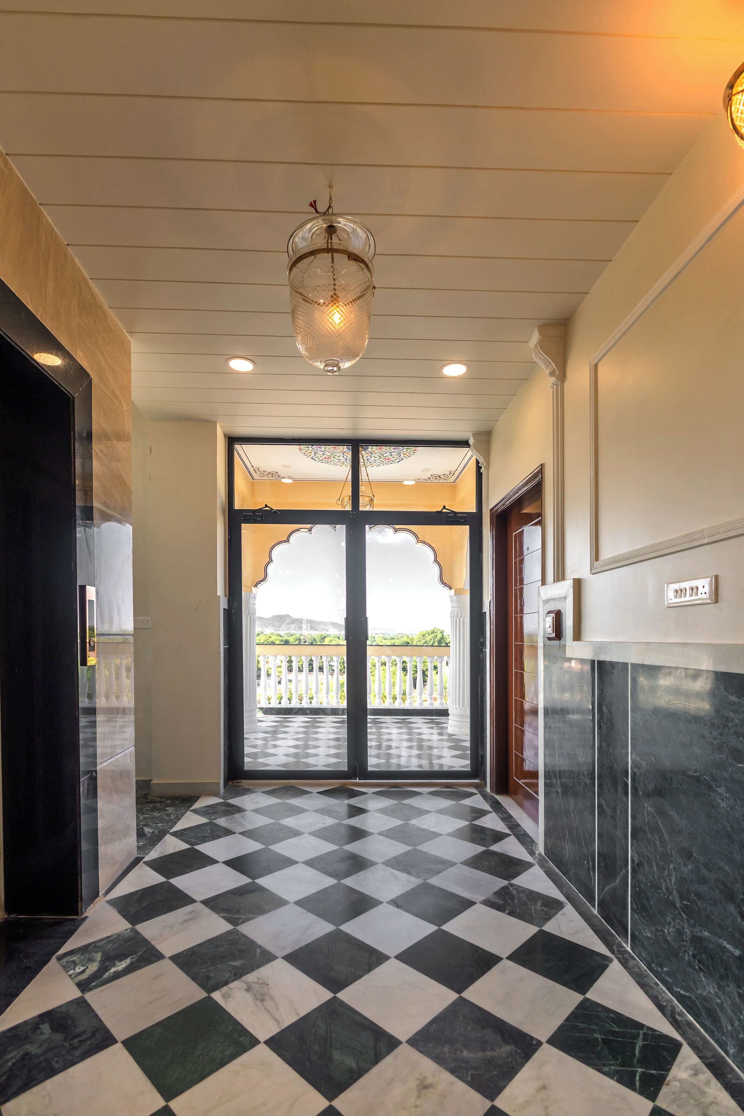 A hotel corridor with a checkered floor, a chandelier, and a view of a balcony with a fence.