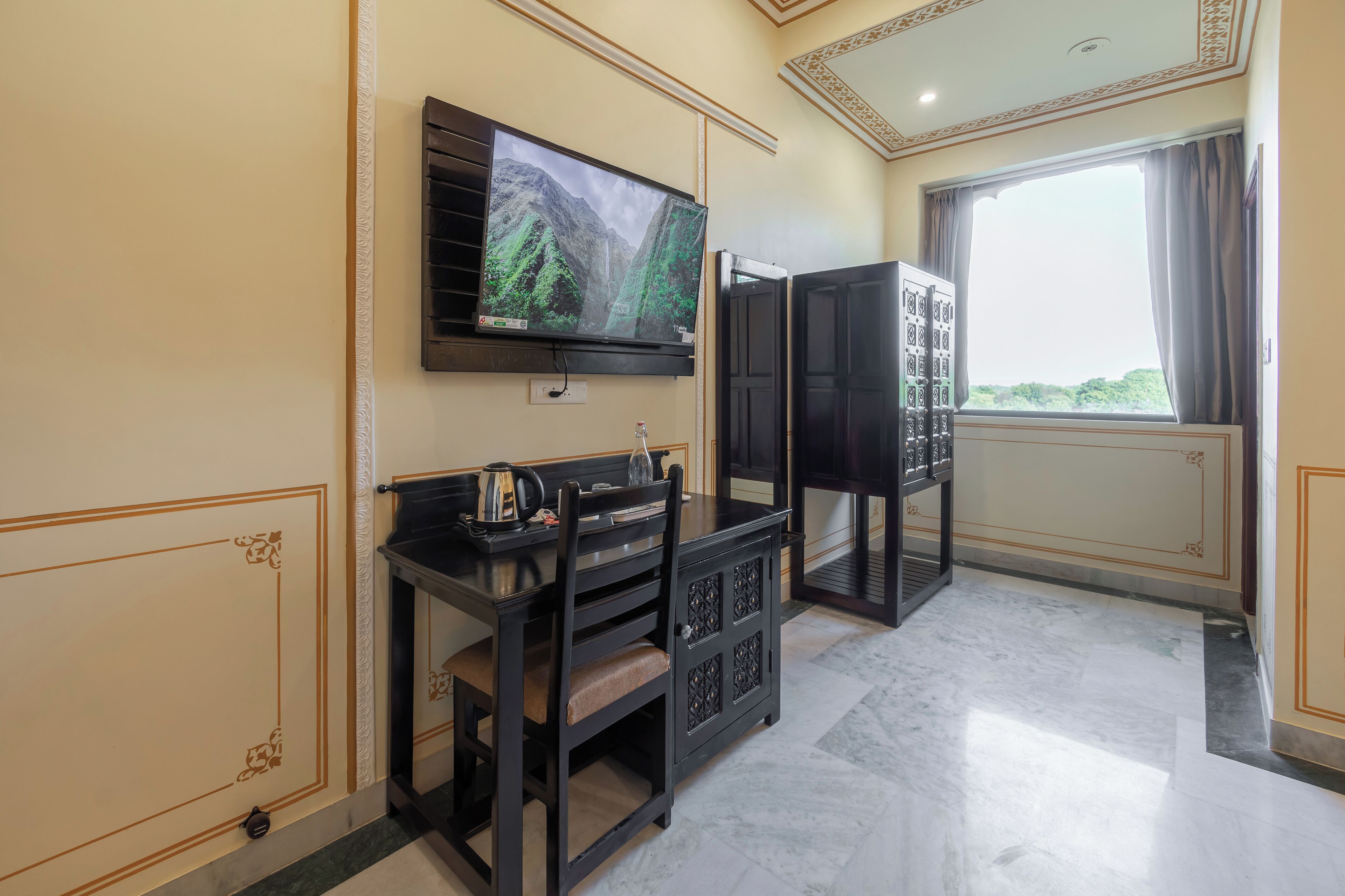 A room with a flat-screen TV mounted on the wall, a dark wooden cabinet with a cushioned seat, and a window with curtains.