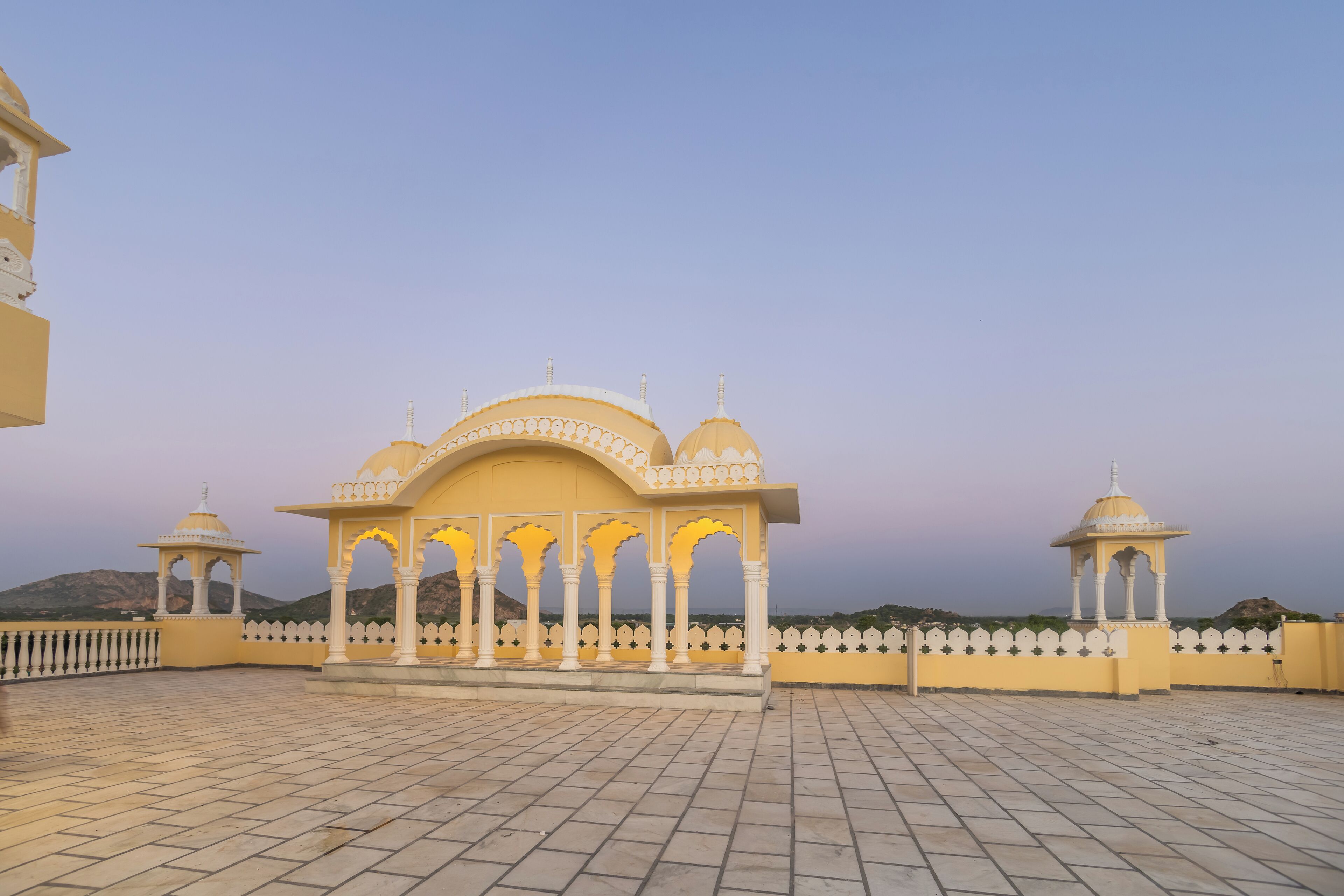 A traditional yellow pavilion with arches and domes, surrounded by a white fence and set against a mountainous backdrop.
