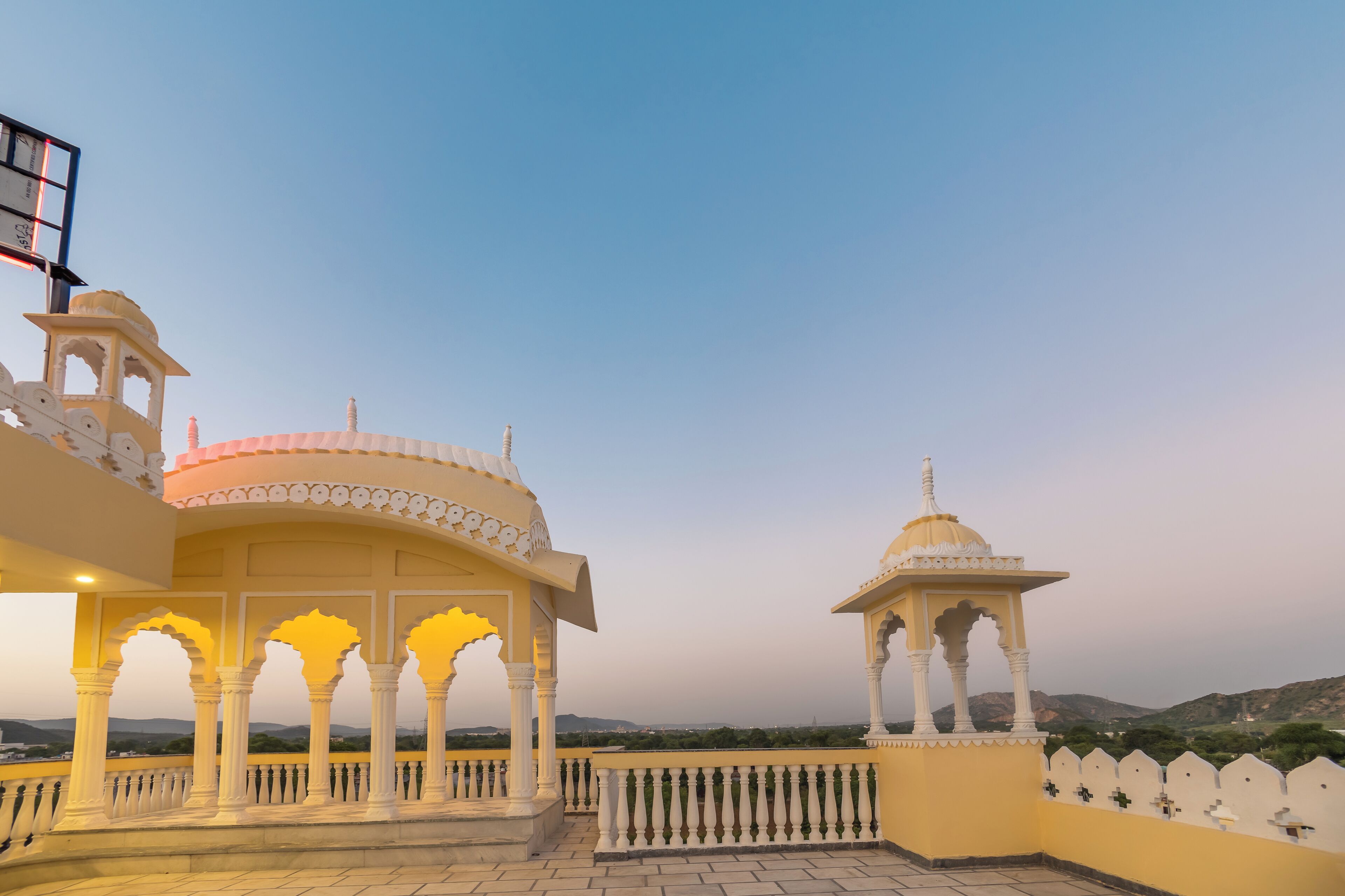 Un pavillon traditionnel doté d’un toit en forme de dôme et de plusieurs arcades, entouré d’une clôture blanche et offrant une vue sur un paysage montagneux.