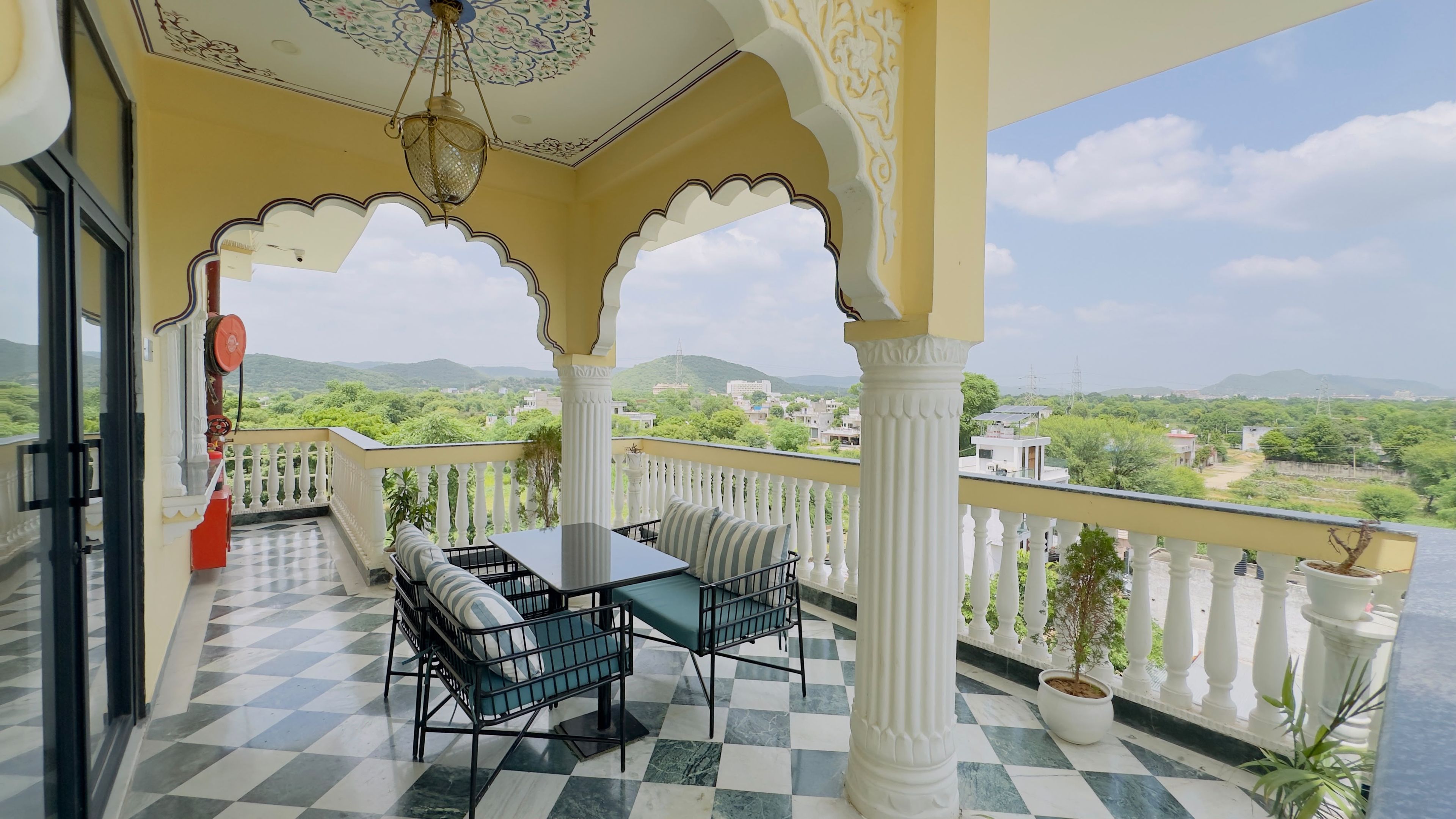 A balcony with a checkered floor, a table with chairs, and a view of greenery and buildings in the distance.