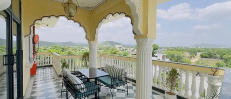 A balcony with a checkered floor, a table with chairs, and a view of greenery and buildings in the distance.