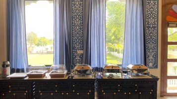 A breakfast buffet setup with covered dishes and a coffee machine, set against a backdrop of blue curtains and a view of a garden.