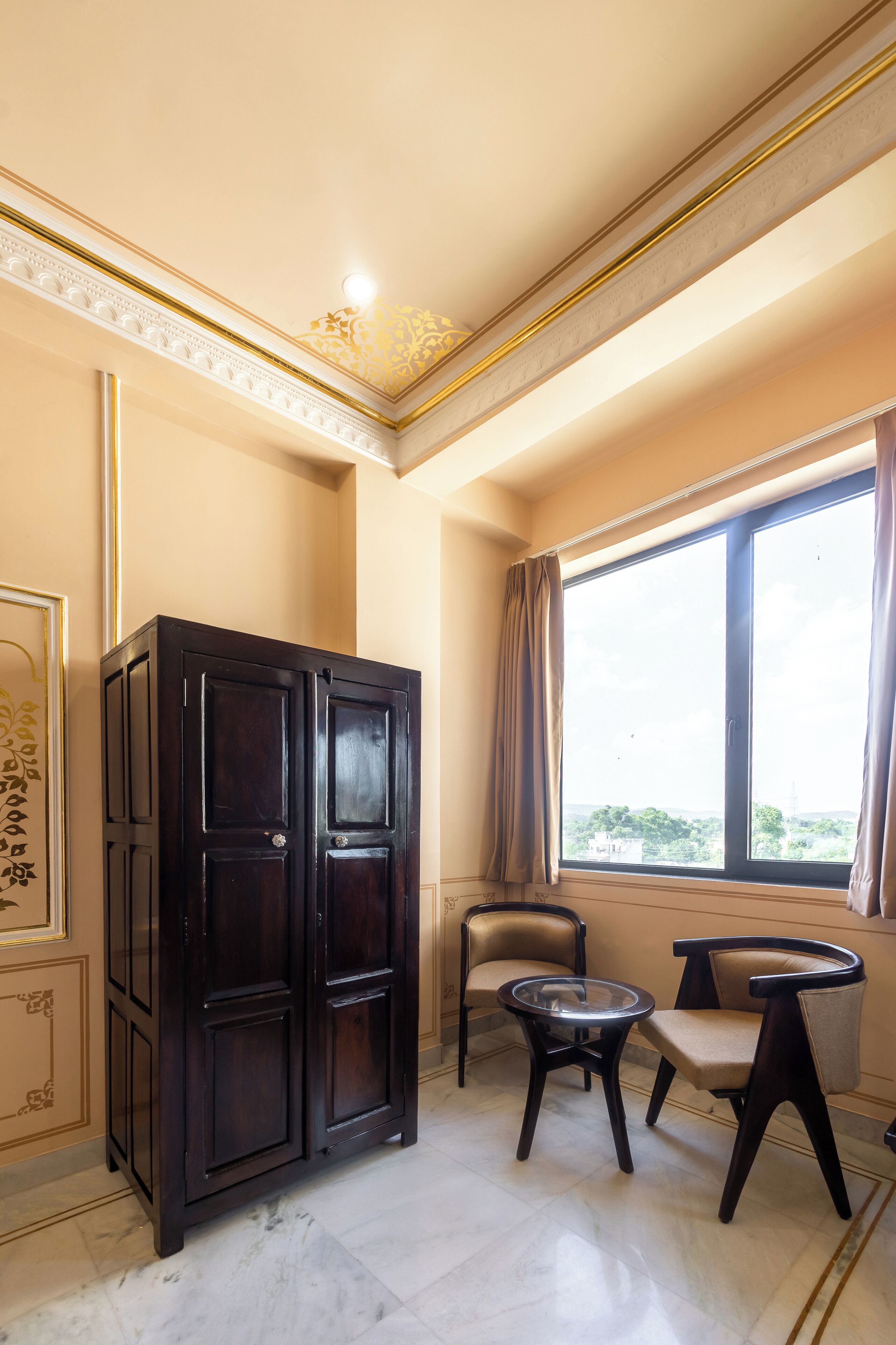 A room with a dark wooden cabinet, two chairs, a small round table, and a large window with curtains.
