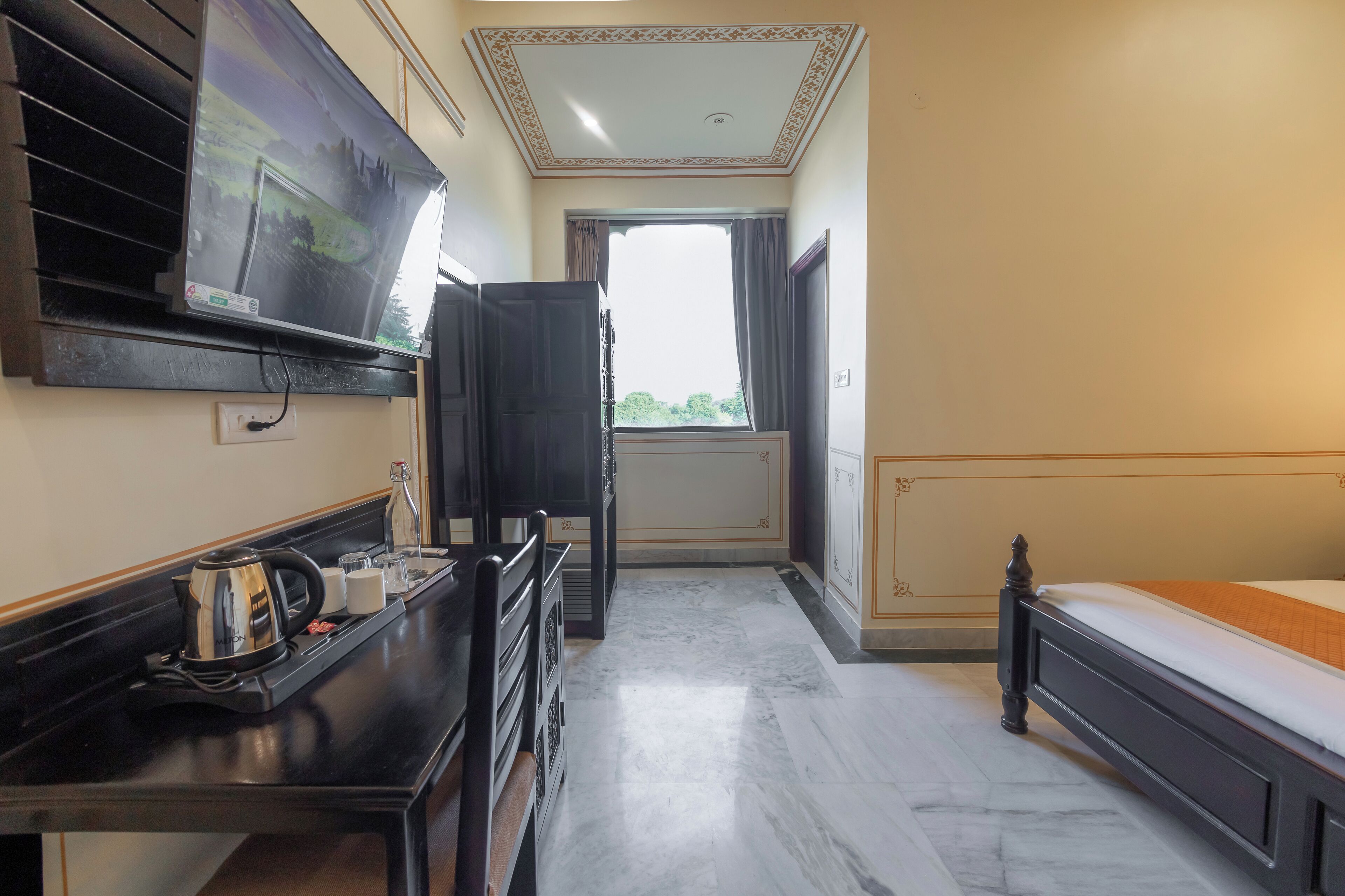 A hotel room with a bed, a TV, a desk with a kettle, and a window with curtains.