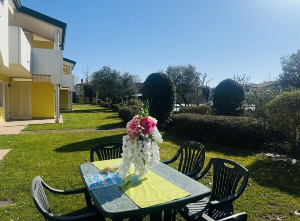 An outdoor dining area with a table set for two, surrounded by chairs. There is a flower arrangement on the table, and the setting includes a well-maintained lawn and a building in the background. - Lovely Flat With Garden (San Michele al Tagliamento)