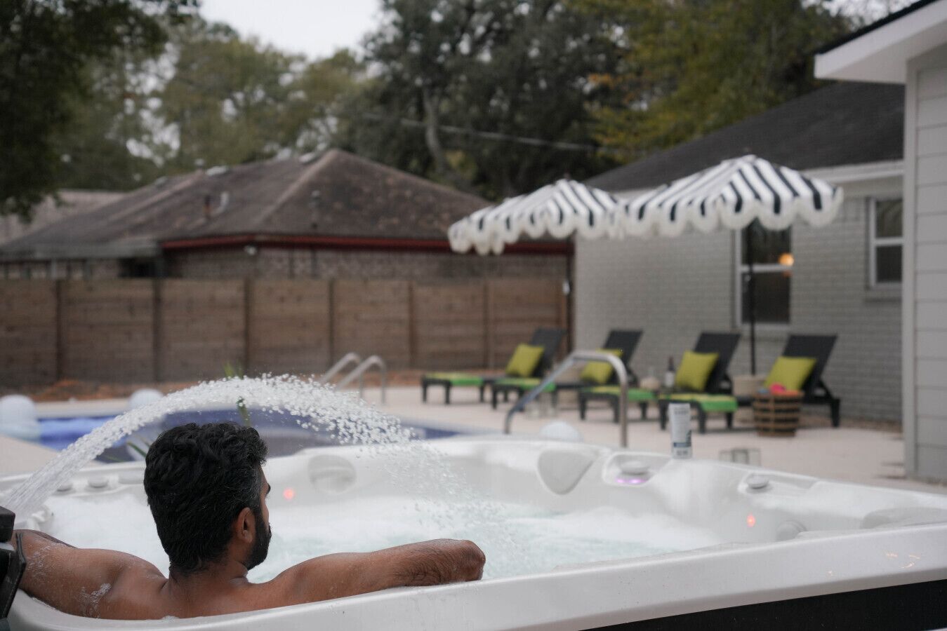 Una persona en un jacuzzi con chorros de agua, rodeado de sillones de exterior y una piscina.
