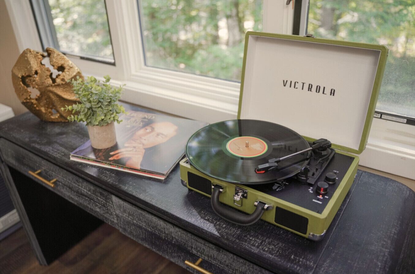 A turntable with a vinyl record on a black table, a green box labeled "VICTROLA," and a small plant.